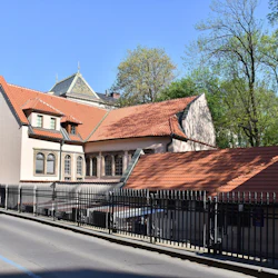 En byggnad med rött tegeltak, välvda fönster och ett svart metallstaket längs en gata med en silverbil framför. Träd i bakgrunden.