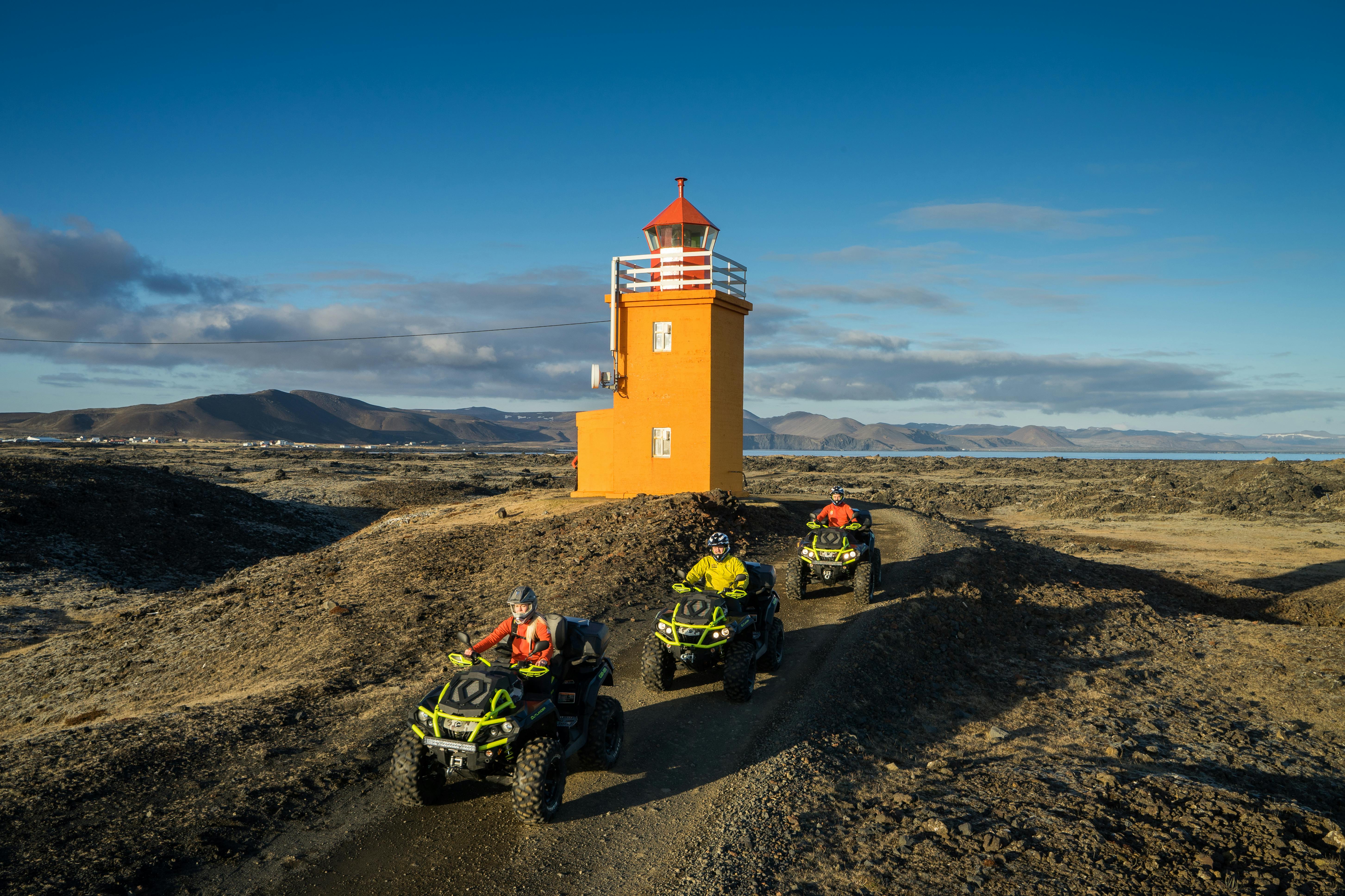 Trois personnes conduisant des VTT sur un chemin de terre à côté d'un phare orange, avec des montagnes et un ciel clair en arrière-plan.