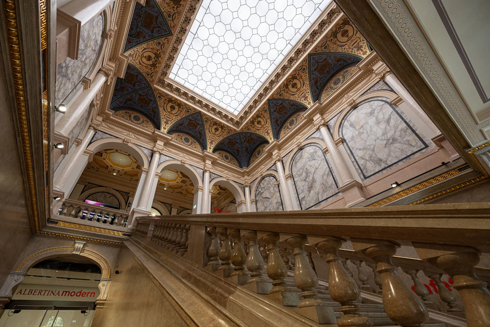 Ornate indoor staircase with marble banisters, vaulted ceiling with intricate designs, and large skylight letting in natural light.