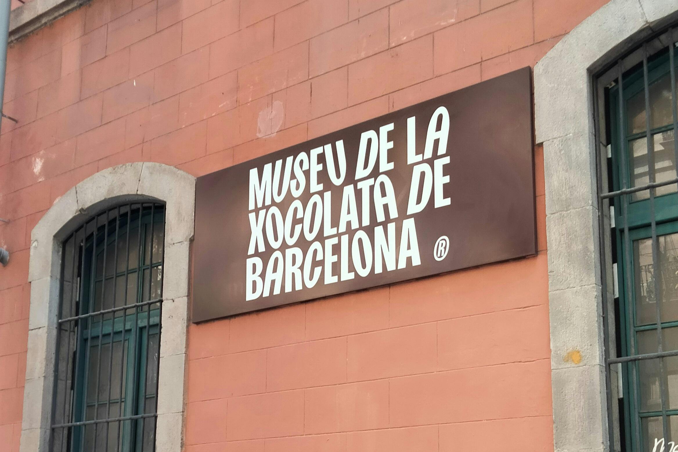 Znak o treści „Museu de la Xocolata de Barcelona” na brązowym tle, zamontowany na budynku o różowawej fasadzie.