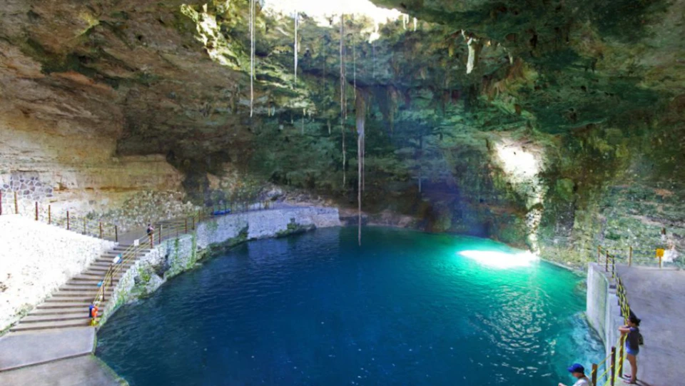 A large underground cave with a pool of clear blue water and hanging stalactites illuminated by sunlight.