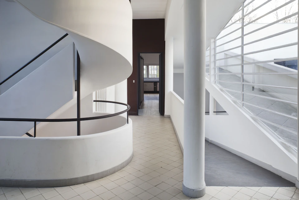 Interior with a white spiral staircase, large windows with horizontal bars, and tiled floor leading to a dark doorway.