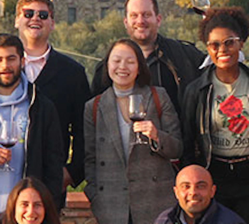 A group of twelve people standing and smiling on a terrace with drinks in hand, scenic countryside and building in the background.