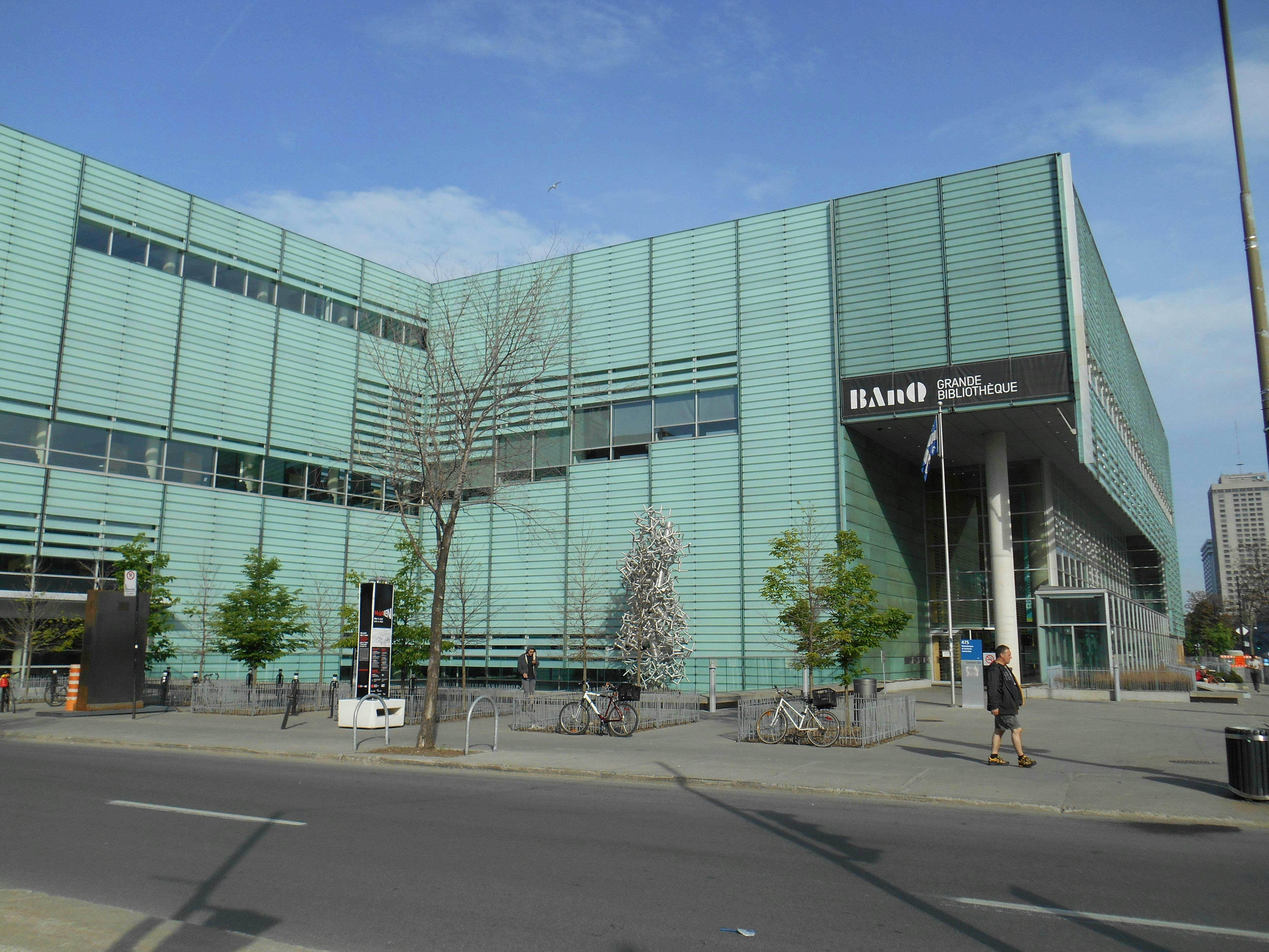 Modern building with teal glass panels, labeled "BAnQ Grande Bibliothèque," bikes parked outside, and a person walking on the sidewalk.
