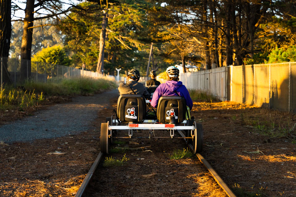 Two people in helmets ride a rail vehicle on tracks surrounded by trees and a fence in a forested area.