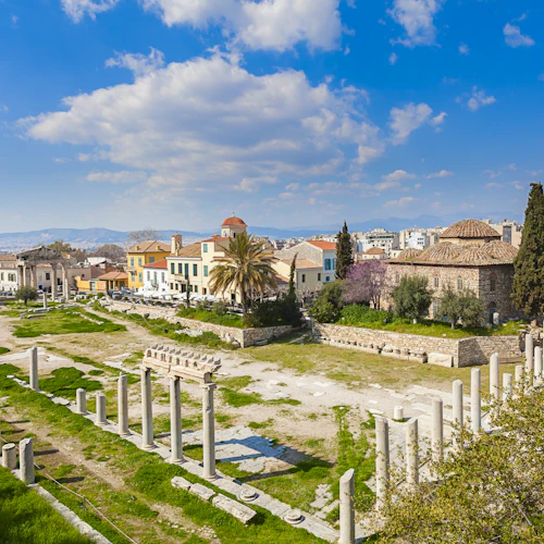 Forntida ruiner med stenkolonner och gräsbevuxna områden, omgivna av historiska byggnader under en blå himmel med spridda moln.