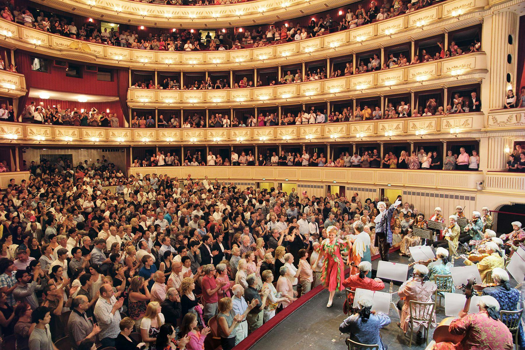 A packed theater with multiple balconies of spectators applauding a performer on stage at the end of a show.