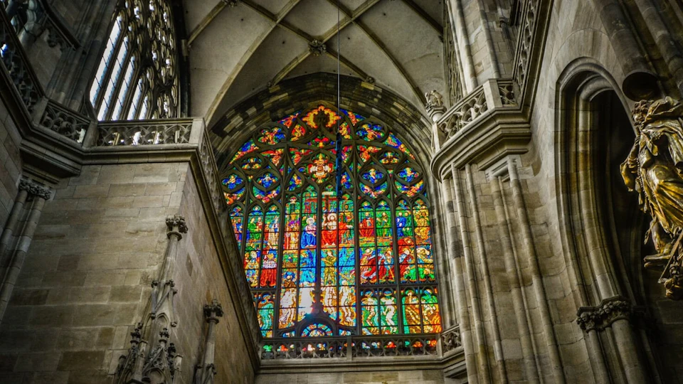 Large stained glass window in a cathedral, depicting vibrant scenes and figures, with arched ceiling and stone architecture.
