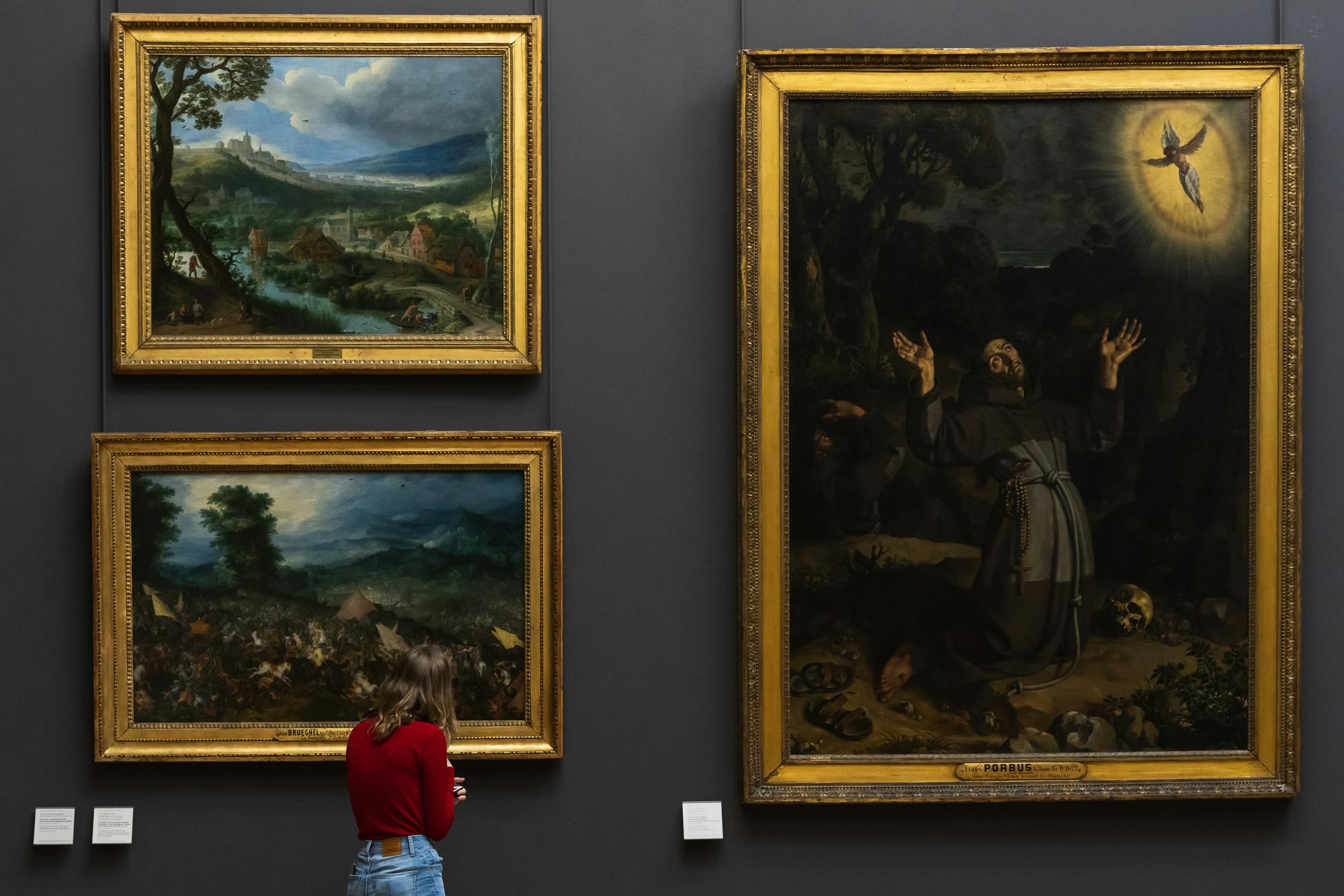 Eine Frau in rotem Hemd und Jeans betrachtet drei gerahmte Gemälde in einer Galerie, darunter zwei kleinere Landschaften und eine große Szene auf der rechten Seite.