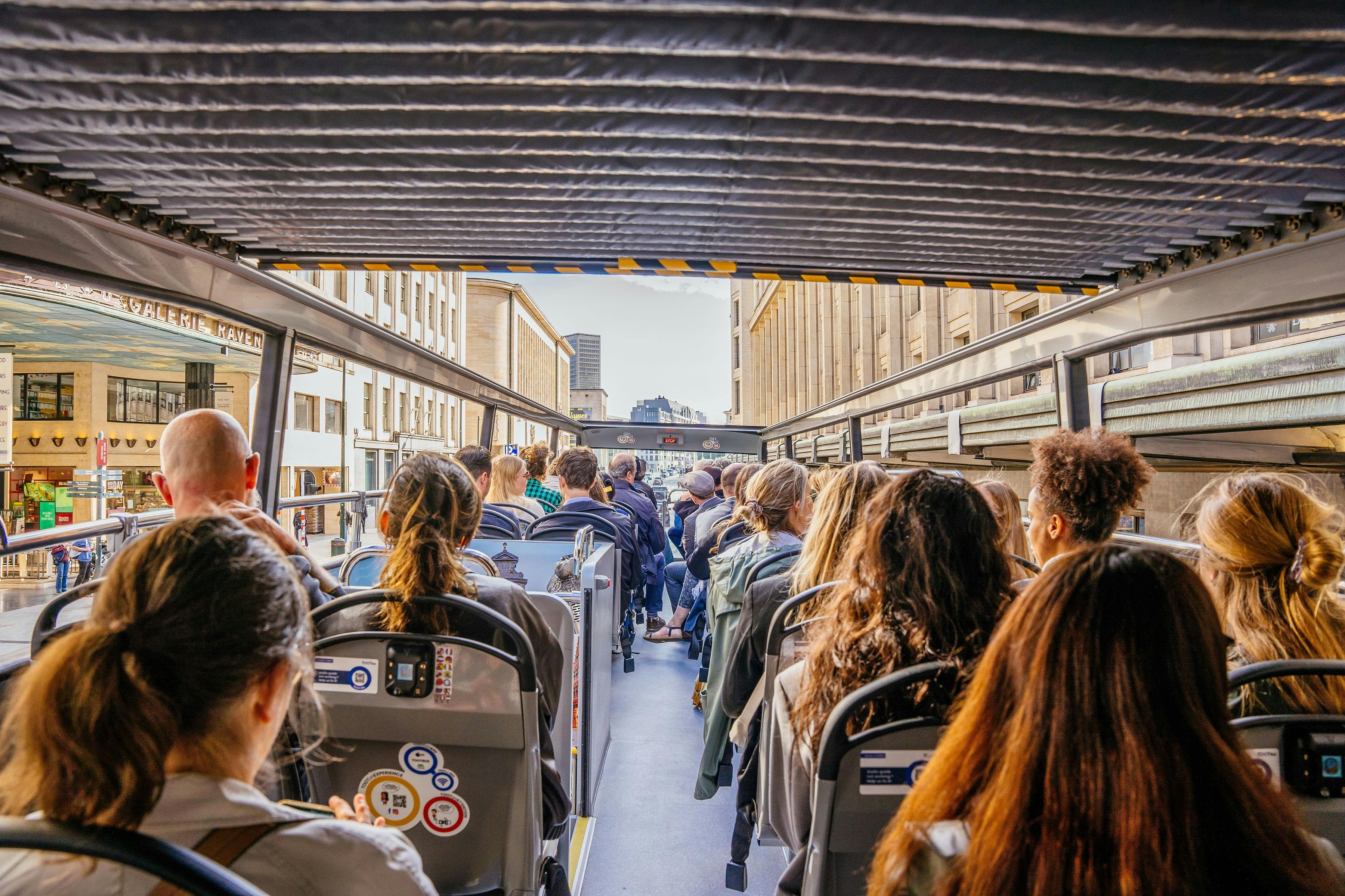 Passengers seated on the upper deck of an open-top tour bus in a city, surrounded by buildings and a clear sky.