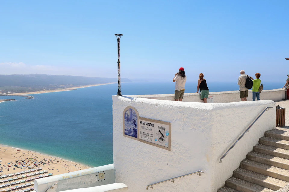 People standing at a white-walled viewpoint, overlooking a beach and ocean. A sign welcomes visitors in multiple languages.
