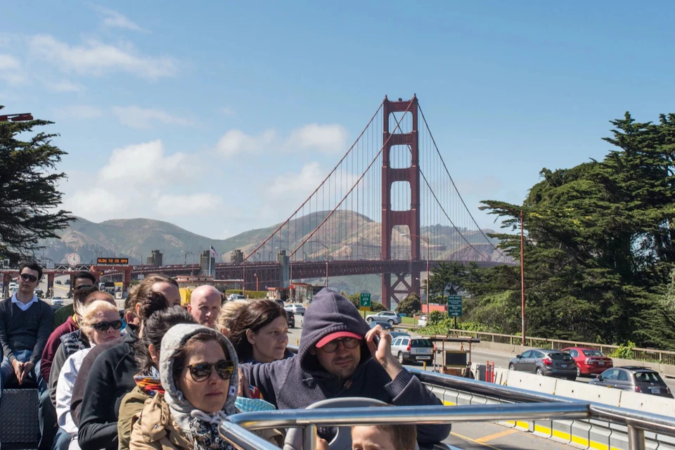People on a tour bus with the Golden Gate Bridge and hills in the background. The sky is clear with scattered clouds.