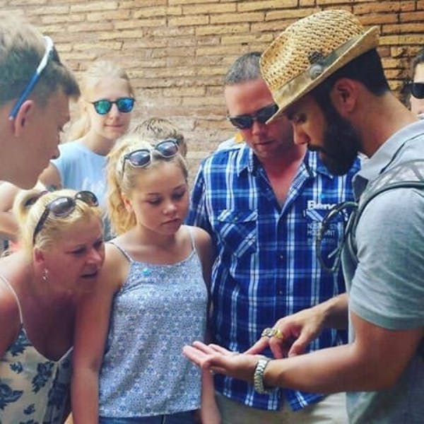 A group of people, including a man in a hat, examines objects in the man's hand. They are standing against a brick wall.