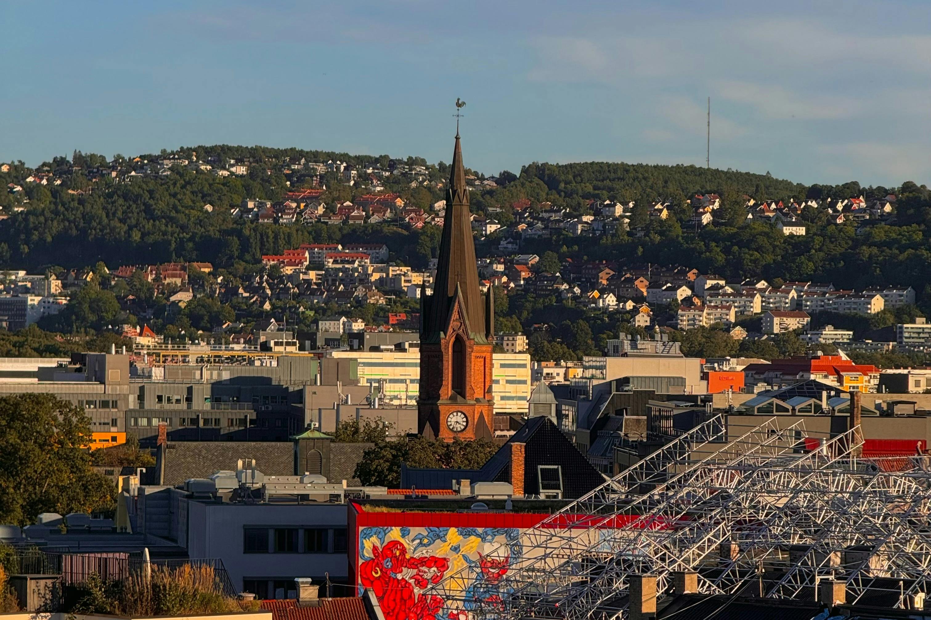 A cityscape featuring a tall church tower with a clock, surrounded by buildings, with wooded hills and houses in the background.