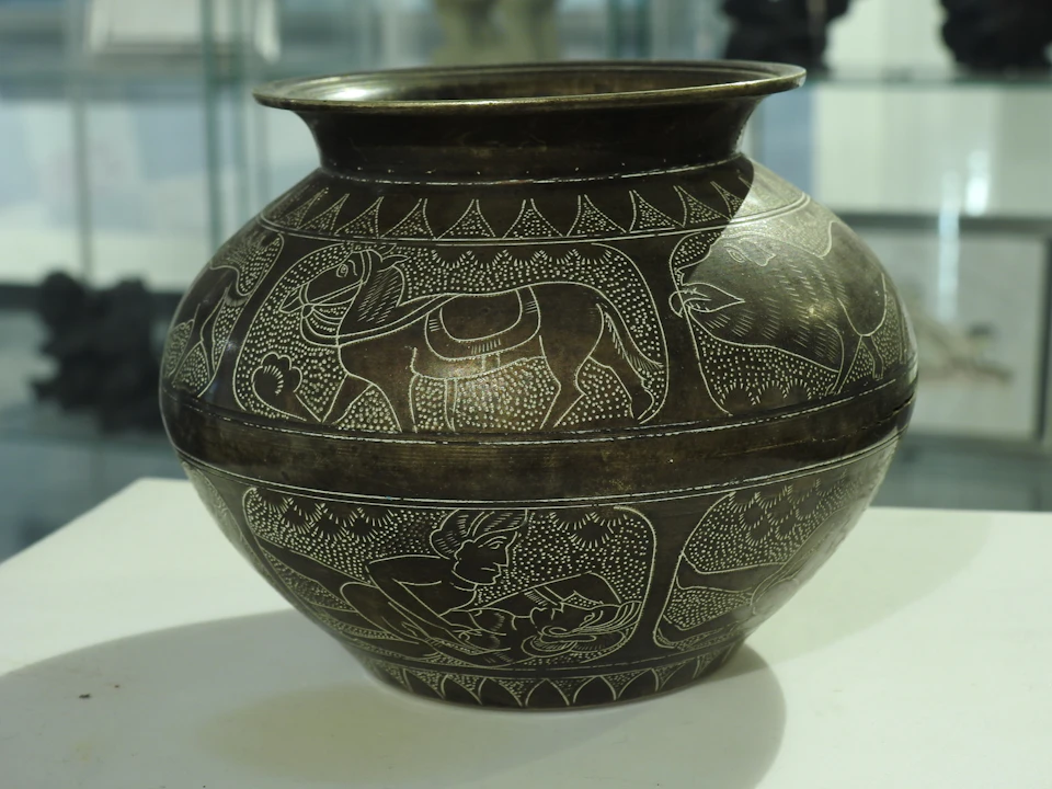 A dark, round pot adorned with white etched animal and human figures, displayed in a glass case.
