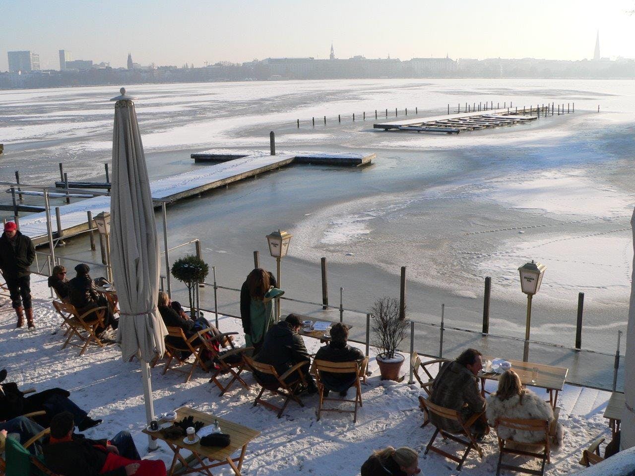 Persone in abiti invernali siedono a tavoli all'aperto su un lungomare ghiacciato e coperto di neve, con moli ed edifici lontani.