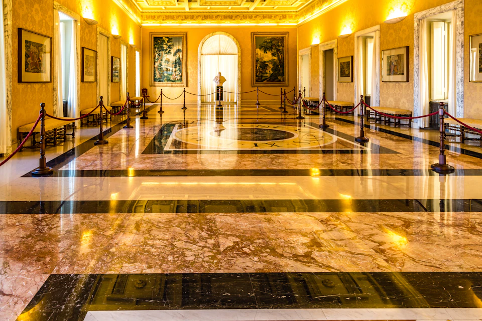Opulent marble-floored hall with artworks on walls, a central medallion, roped-off areas, and a person standing by an arched window.
