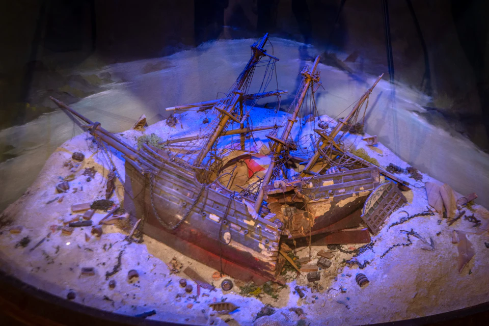 Model of a shipwreck on a sandy sea floor, illuminated with blue lighting. Broken masts and scattered debris are visible.