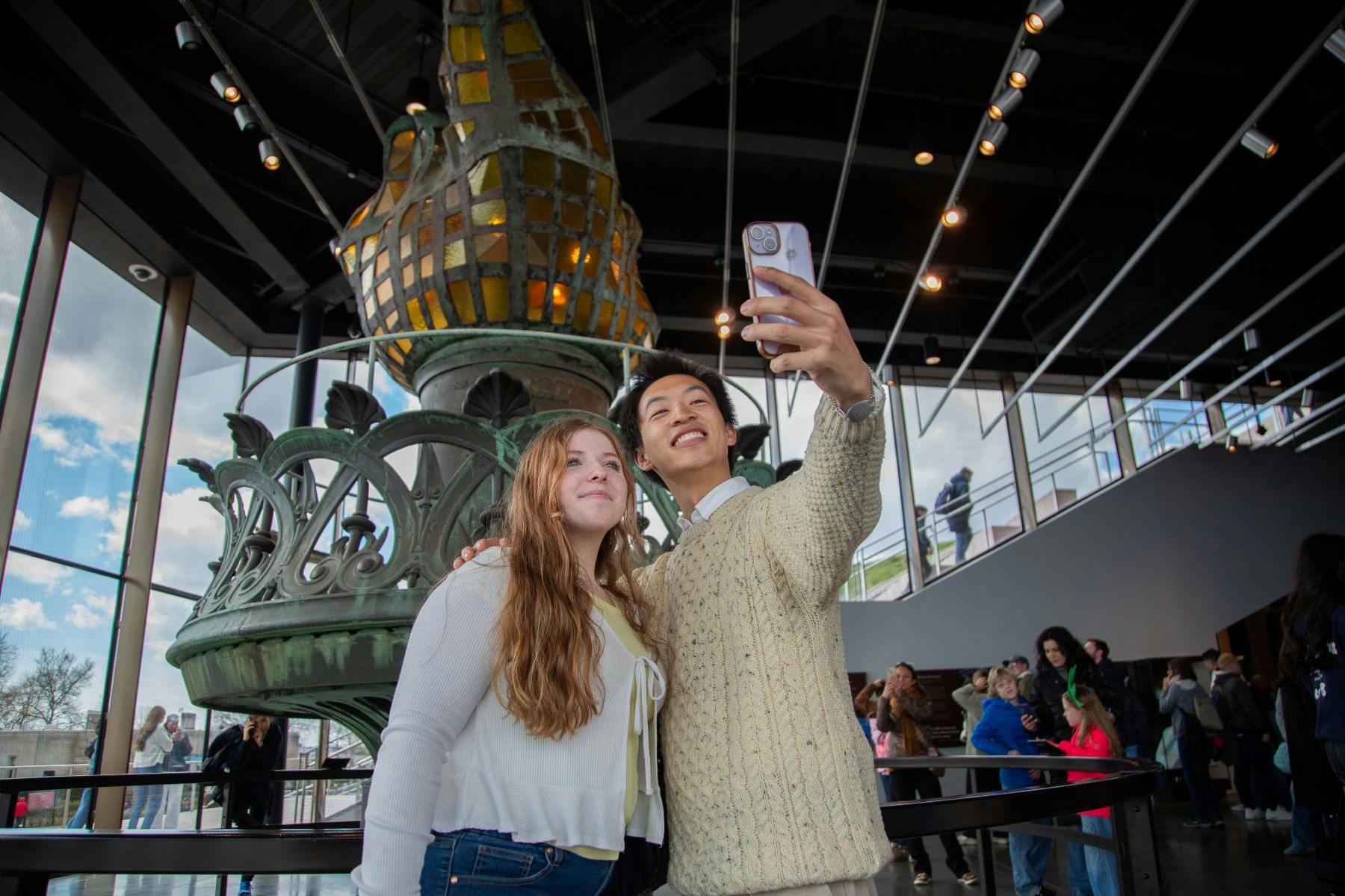 Twee mensen nemen een selfie voor een groot decoratief object in een modern gebouw met een menigte op de achtergrond.
