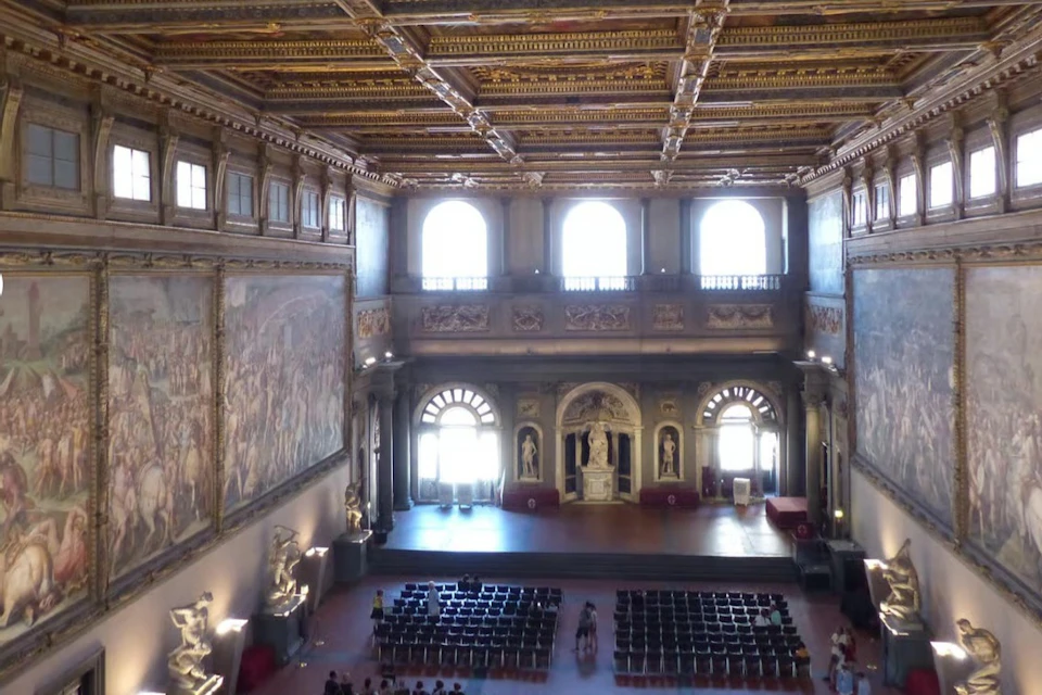 A grand hall with ornate ceiling, large arched windows, statues, wall murals, rows of chairs, and a stage area.