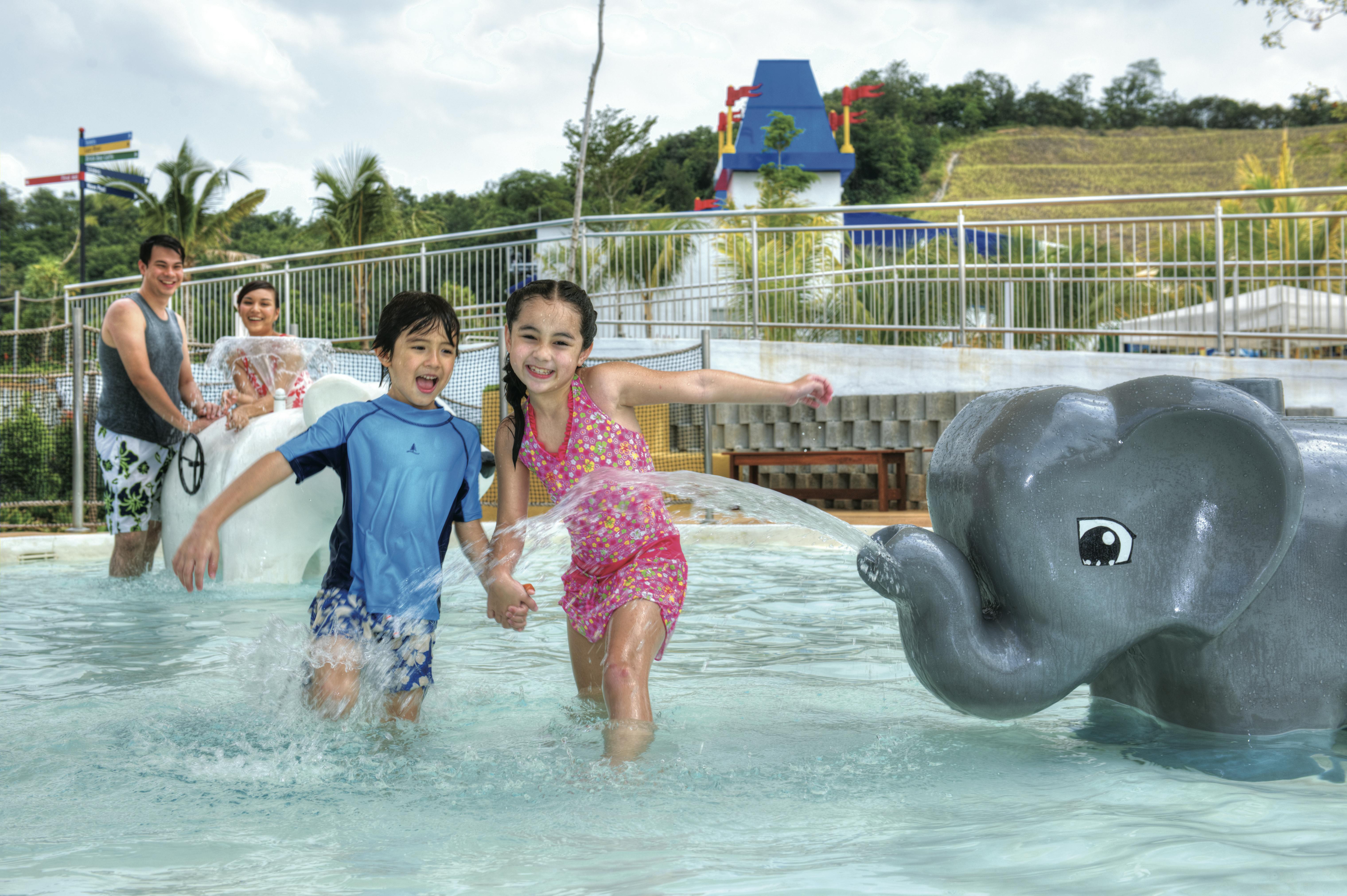Two children play and splash in a shallow pool, while two adults look on smiling. Tropical background with slides and attractions.