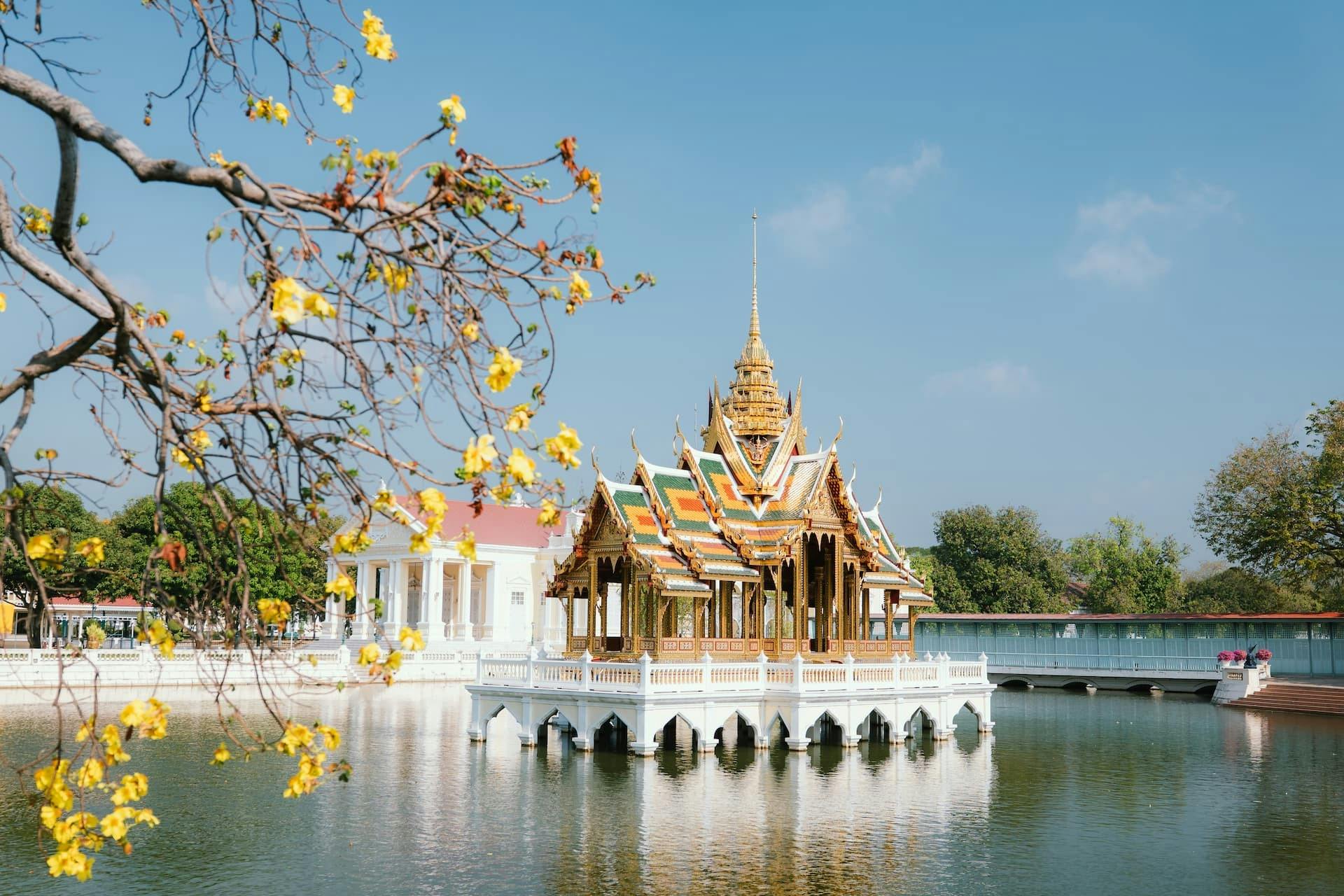 En traditionel thailandsk pavillon på en reflekterende dam, omgivet af træer og en hvid bygning under en klarblå himmel.