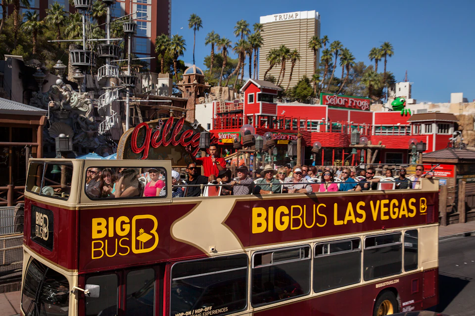 Tourists sitting on an open-top Big Bus tour bus in Las Vegas, with casinos, palm trees, and neon signs in the background.