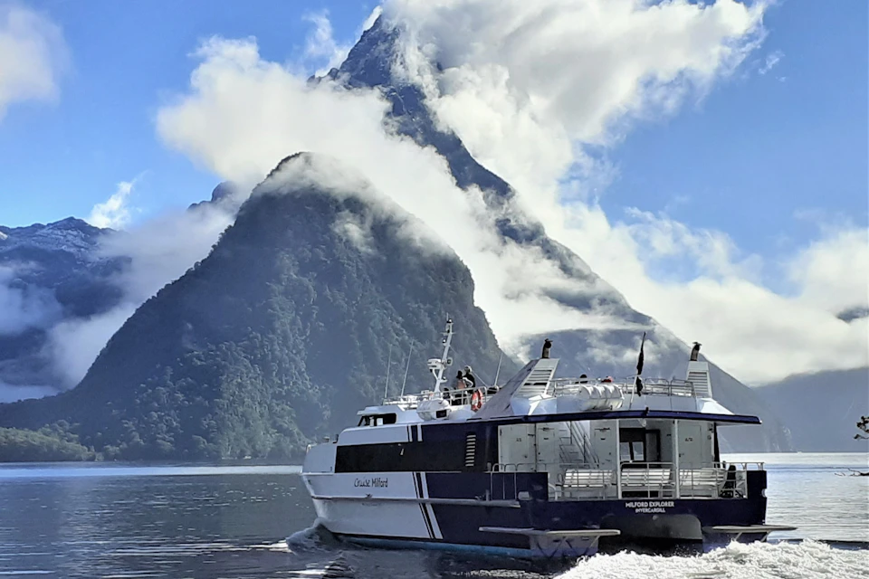 A boat on calm water with a steep, cloud-covered mountain in the background under a blue sky.