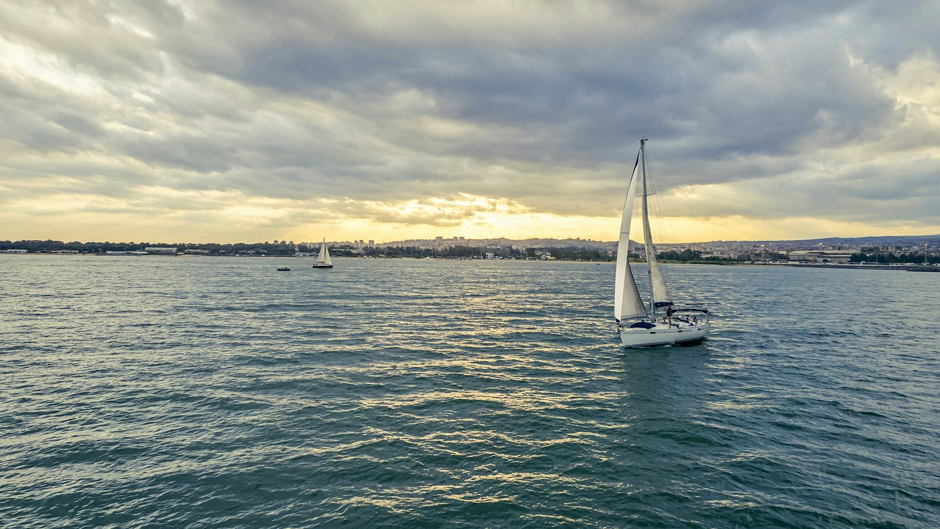 Dvě plachetnice na klidné vodní hladině pod zataženou oblohou se vzdáleným pobřežím viditelným v pozadí.