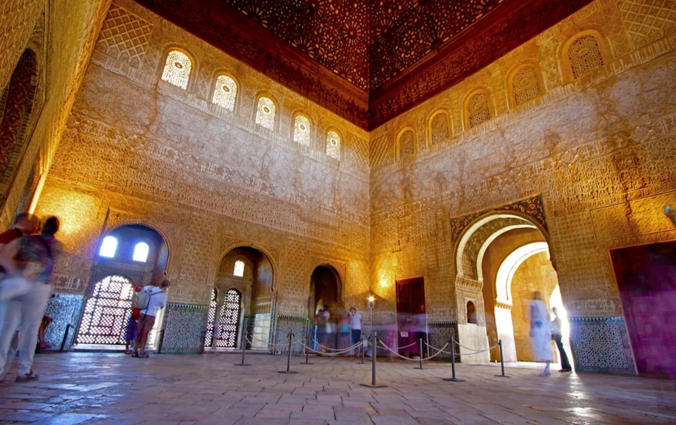 Interior of a historic building with intricate wall carvings, arched windows, and visitors walking by.