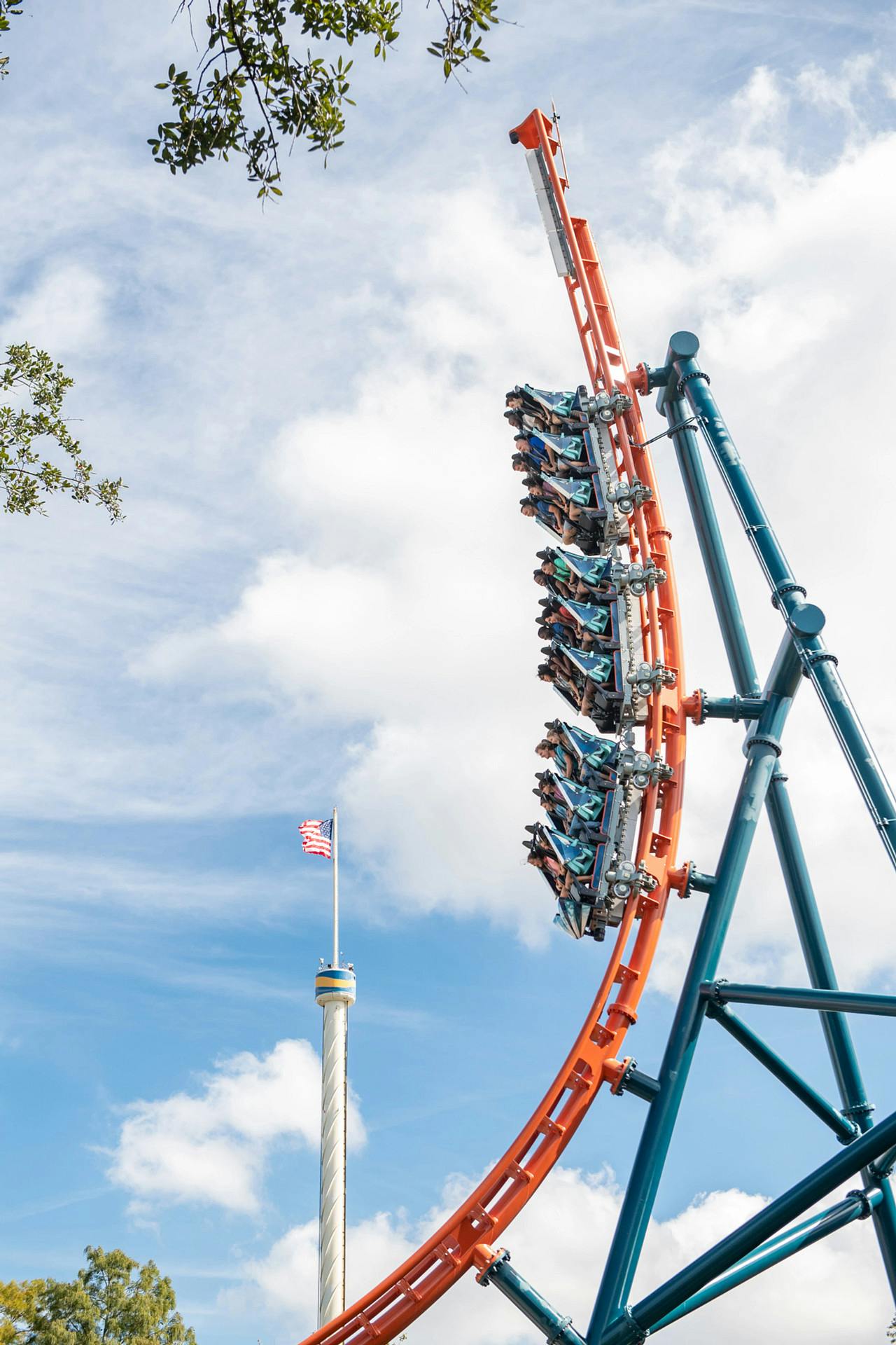 人们坐在陡峭的过山车上，面对多云的天空弯曲，背景中可以看到旗杆和塔楼。