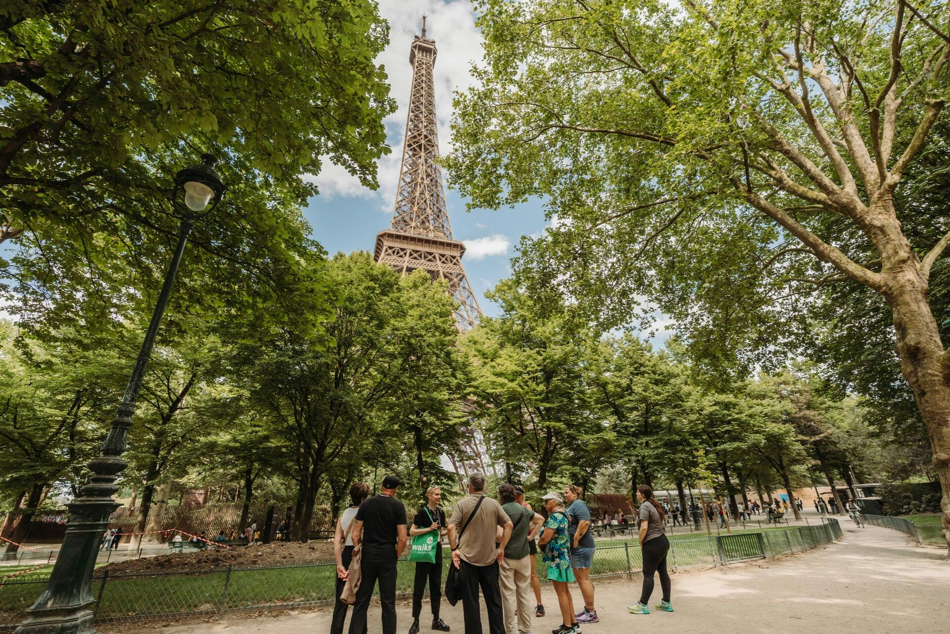 A group of people stand in a park with the Eiffel Tower visible behind trees on a sunny day.