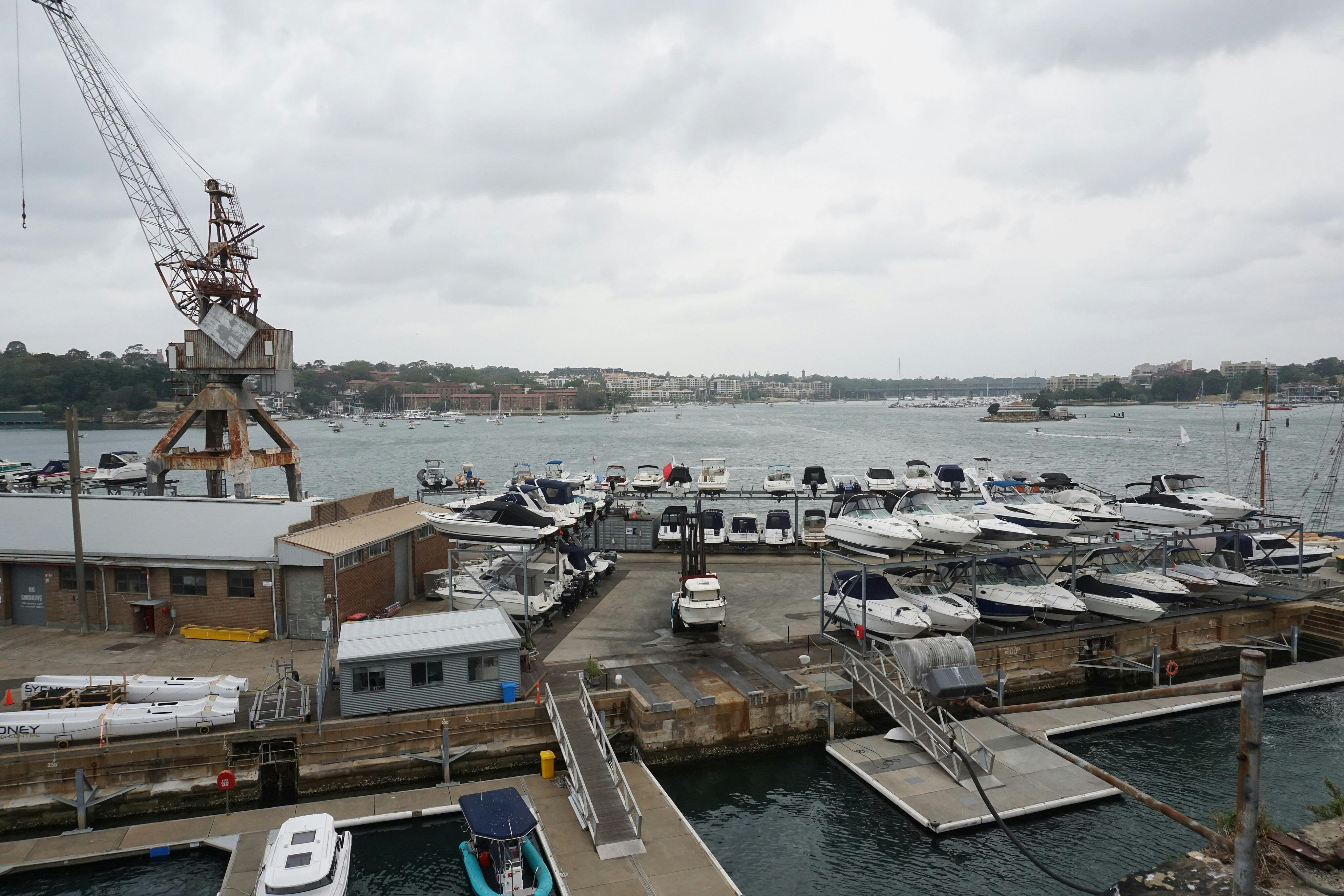 ボートが係留され積み上げられた造船所、古いクレーン、ウォーターフロントの建物、遠く離れた住宅地に広がる曇り空。