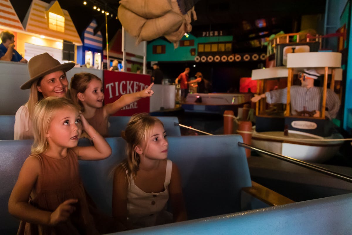 一个有三个小女孩和一个成年人的家庭坐在游艇上，兴奋地凝视着一个明亮、色彩缤纷的游乐园里的场景。