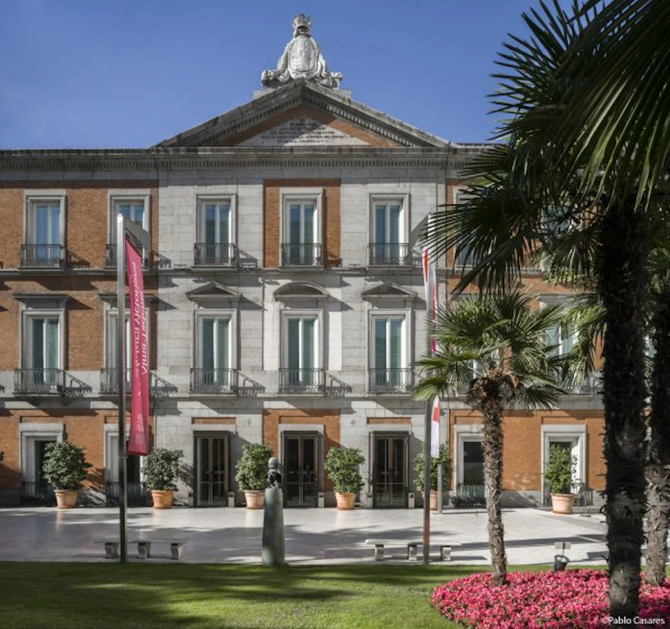 Ornate building with columns and large windows, surrounded by palm trees, pink flowers, and red banners on a sunny day.