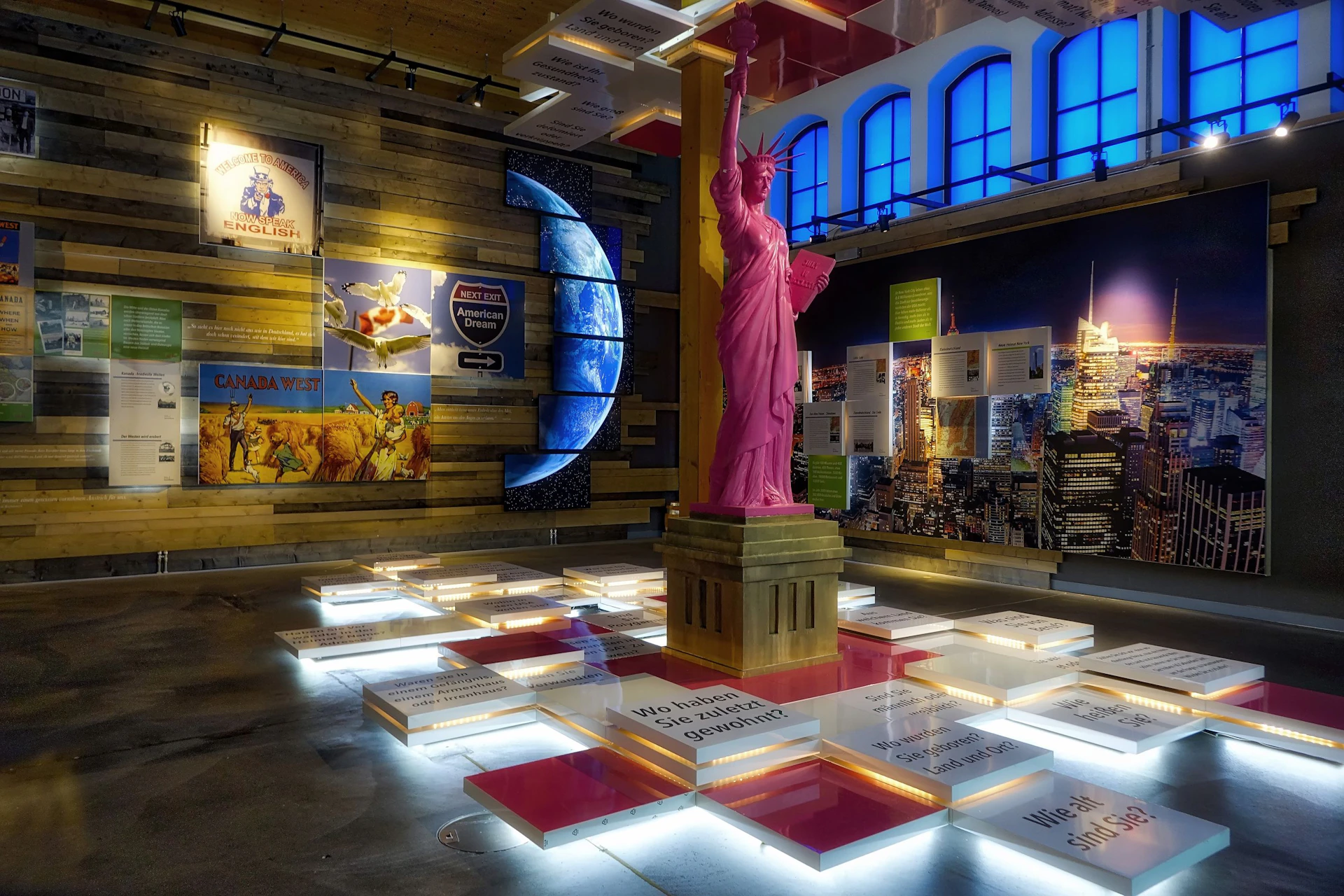 Indoor exhibit featuring a pink Statue of Liberty replica surrounded by illuminated display panels and various wall posters.