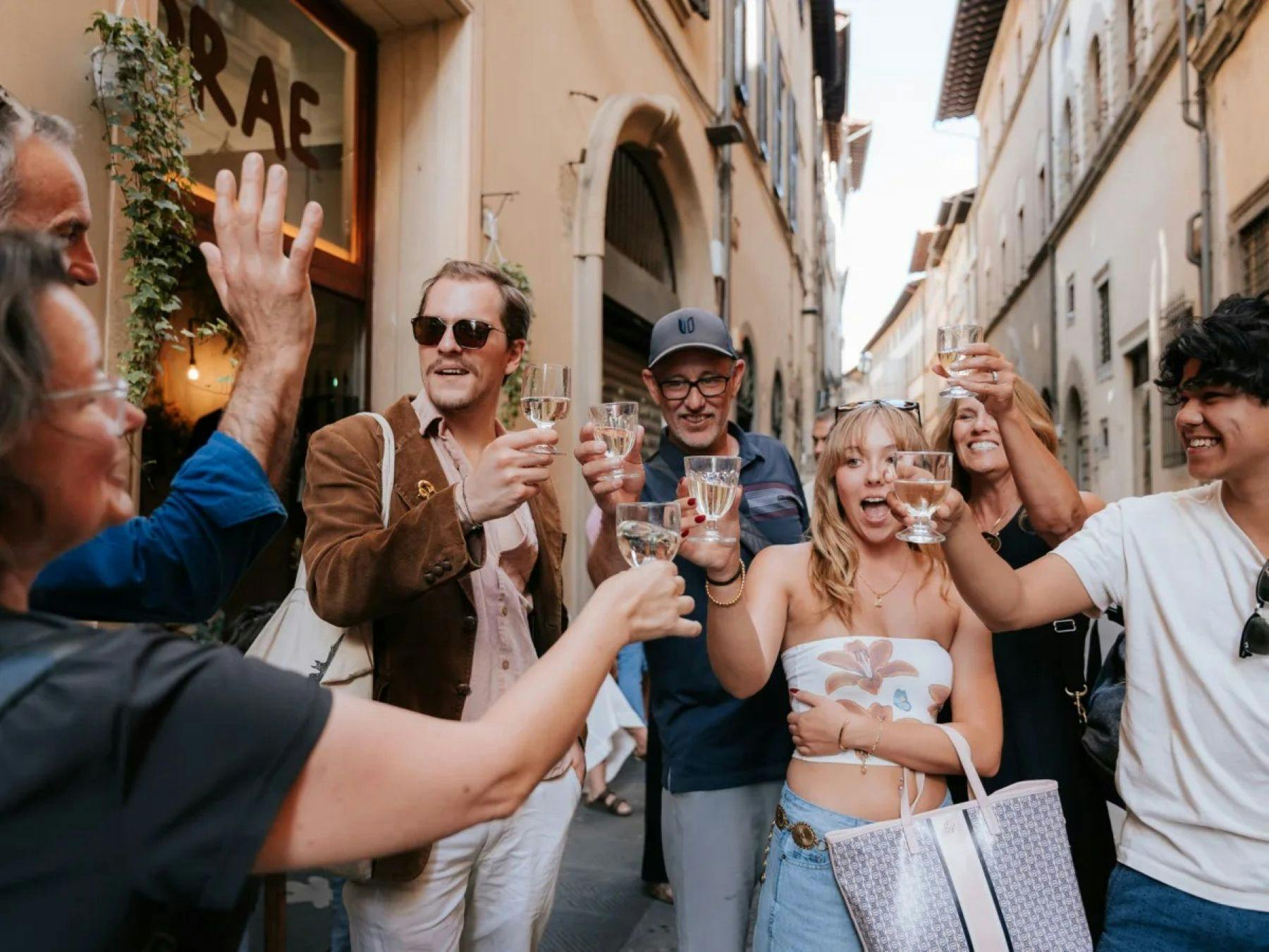 Un gruppo di persone che brindano allegramente con dei bicchieri di vino in una strada soleggiata in un contesto urbano.