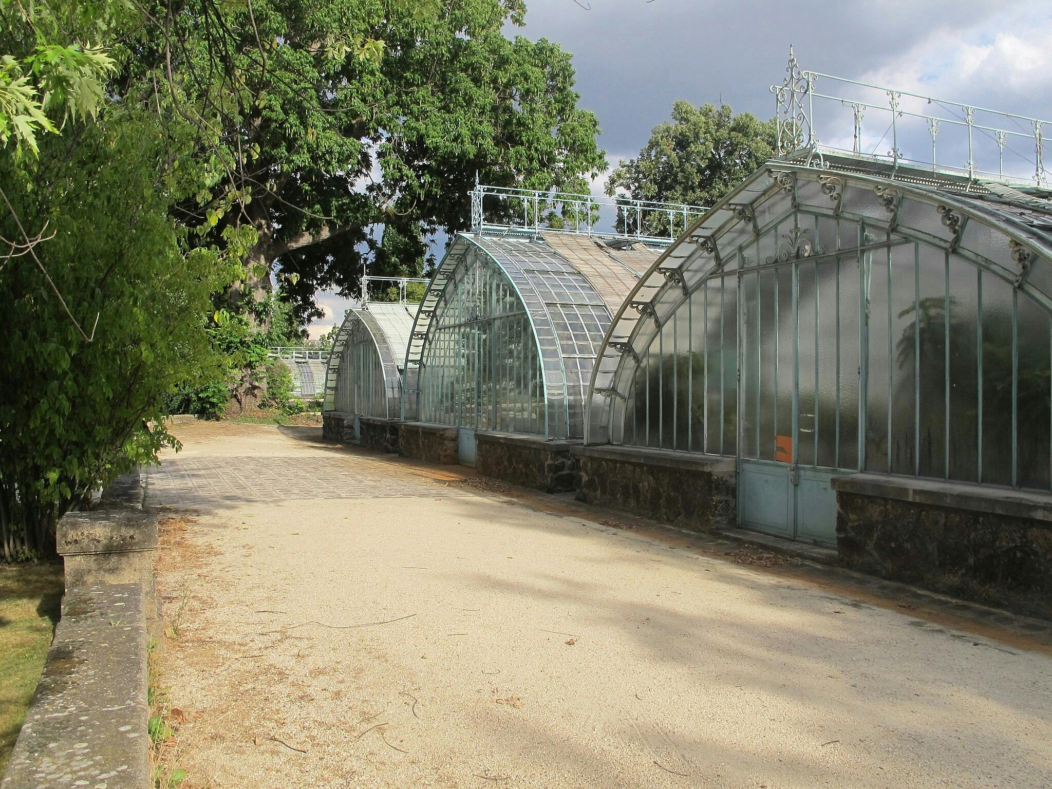 Un sentier mène à de grandes serres en verre incurvées, entourées de verdure et d'arbres, sous un ciel partiellement nuageux.