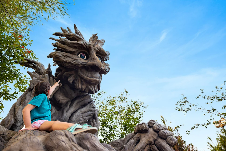 A child interacts with a large, whimsical tree-like sculpture under a bright blue sky with some greenery in the background.