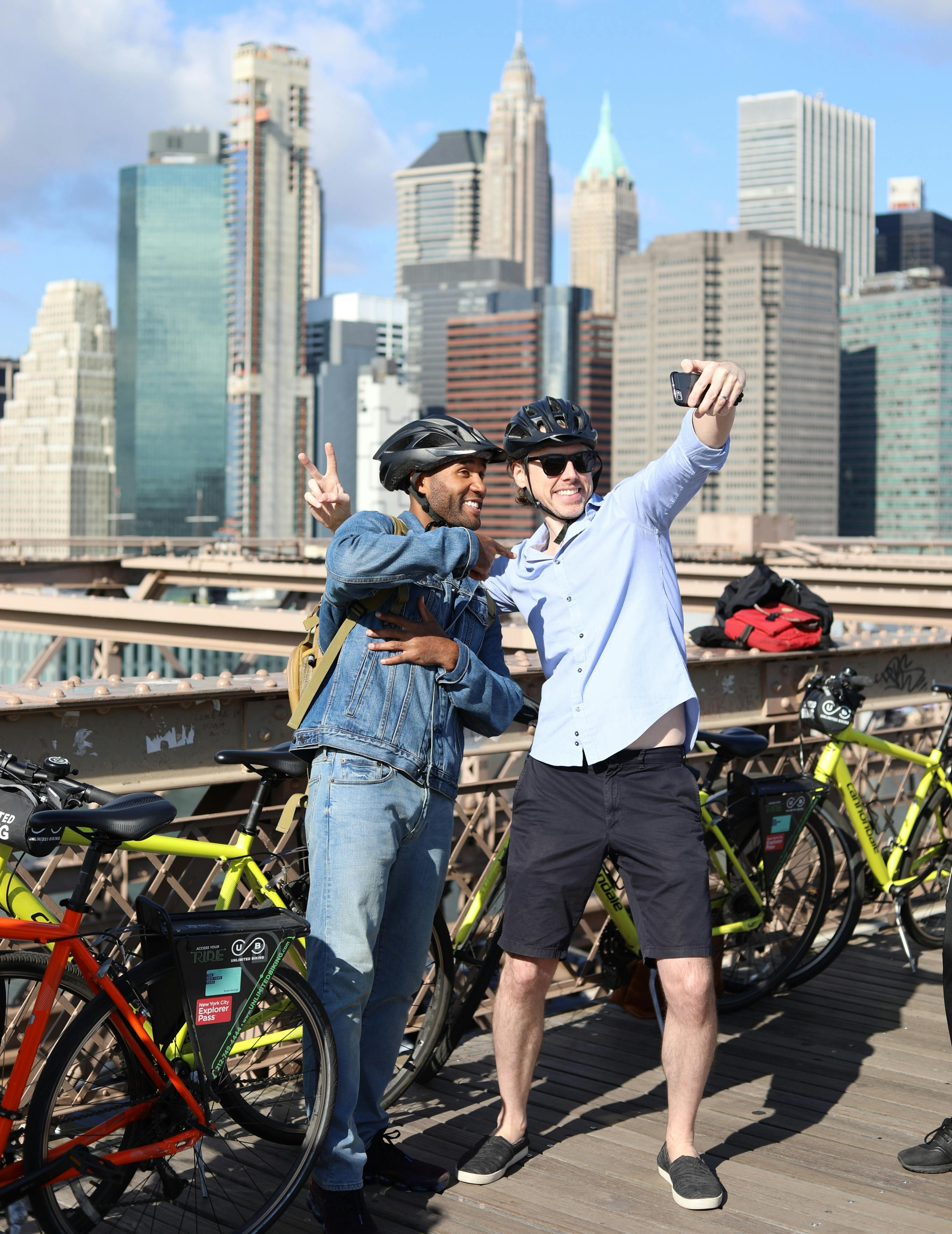 Due persone con il casco si scattano un selfie su un ponte con biciclette, grattacieli e un cielo limpido sullo sfondo.