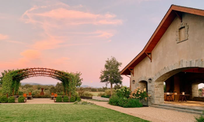 Okouzlující venkovská scéna s velkým domem, zahradním obloukem, lavičkami, stromy a rozlehlým trávníkem pod pastelovým západem slunce.