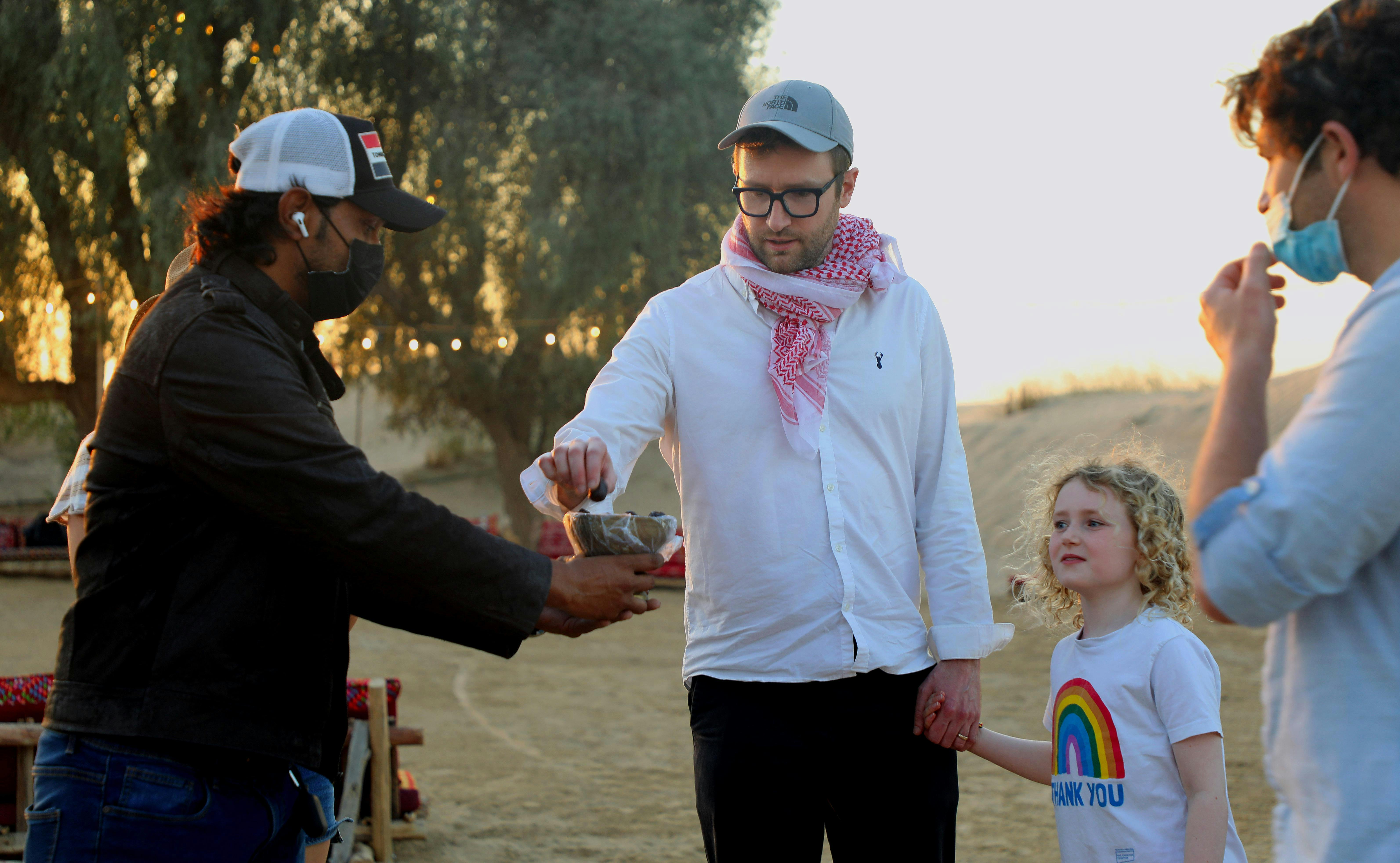 穿着白色衬衫和围巾的男子一边喝酒，一边握着女孩的手。 另一名男子戴着口罩。 背景是沙丘和树木。