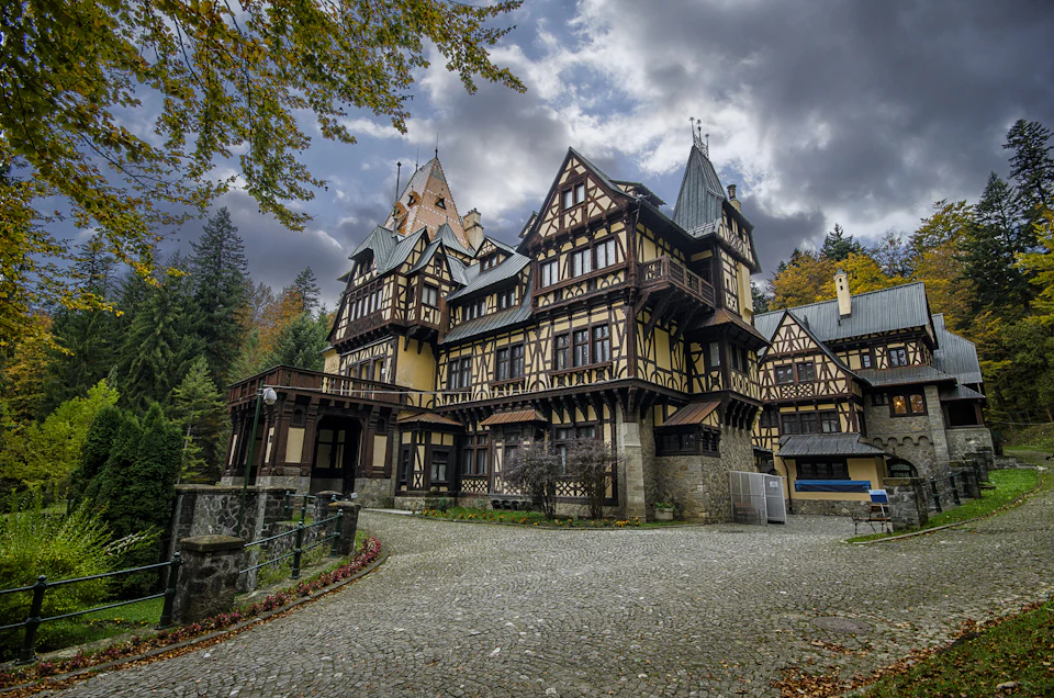 Ornate, multi-story timber-framed building with steep roofs and decorative woodwork, set in a wooded area under a cloudy sky.