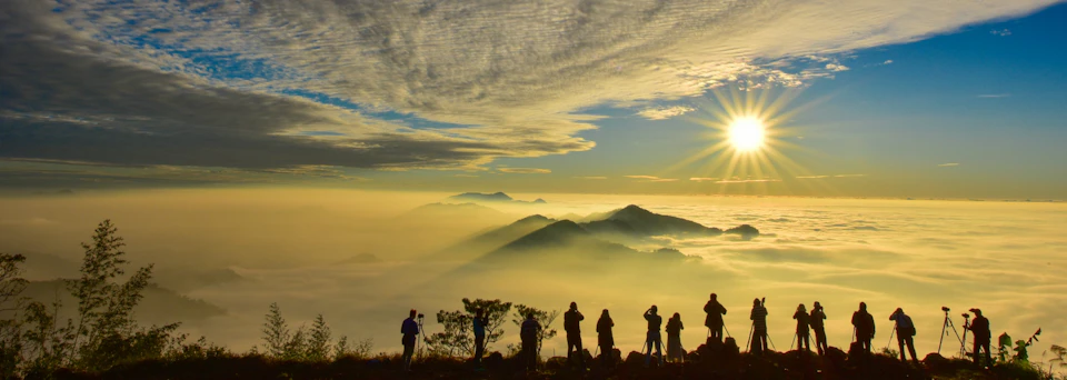 Silhouetted people observe a misty mountain landscape under a vibrant sunrise with scattered clouds.