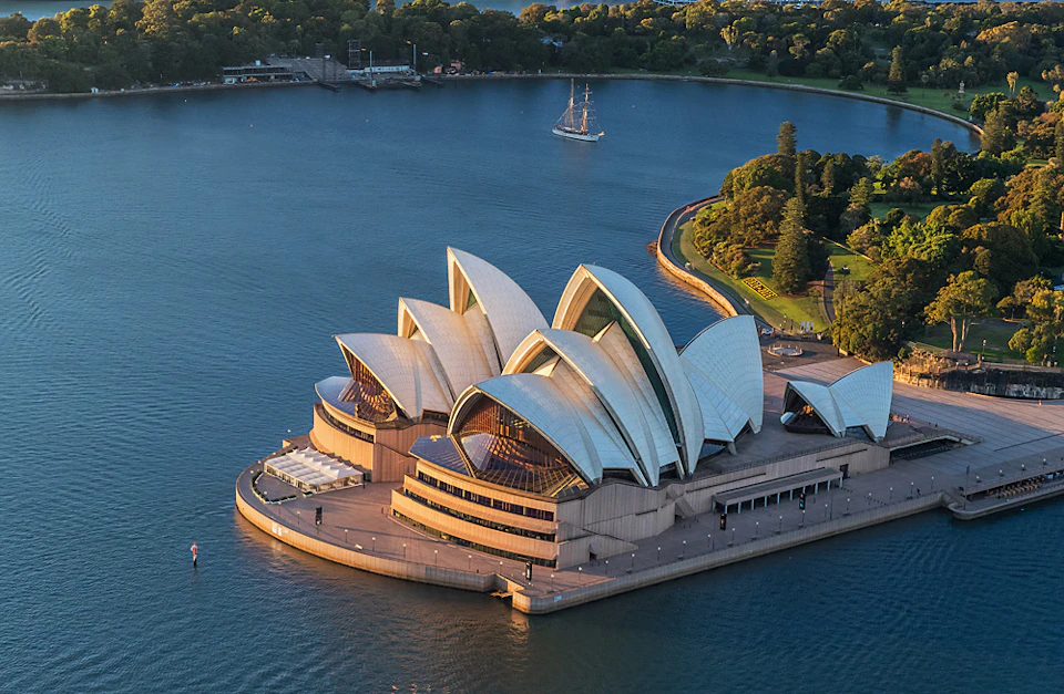 Aerial view of the Sydney Opera House with its iconic white sails, surrounded by water and greenery, and a sailboat nearby.