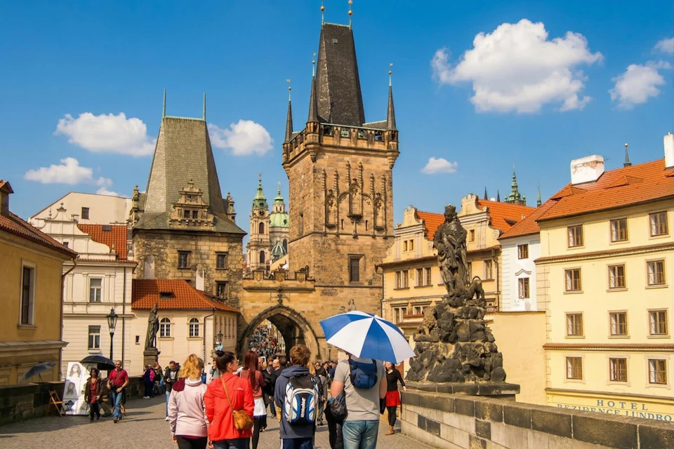 A bustling European street with historic architecture, a tall gothic tower, and tourists walking under a clear blue sky.