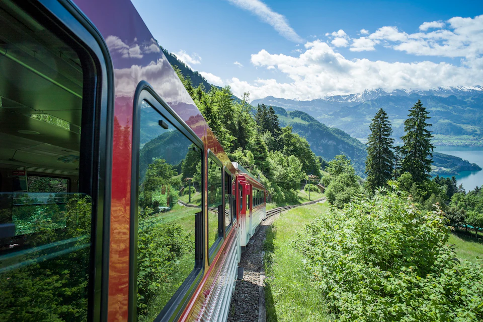 A red and white train travels through a lush, green mountain landscape on a sunny day, with snow-capped mountains in the distance.