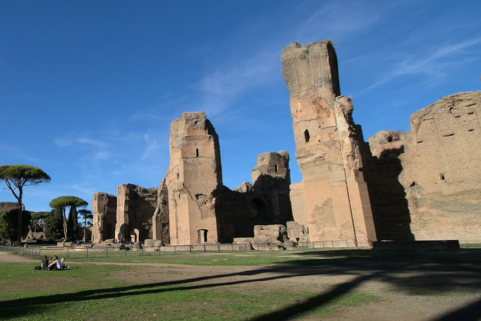 Ancient brick ruins of tall, weathered structures under a clear blue sky, with green grass and shadows in the foreground.