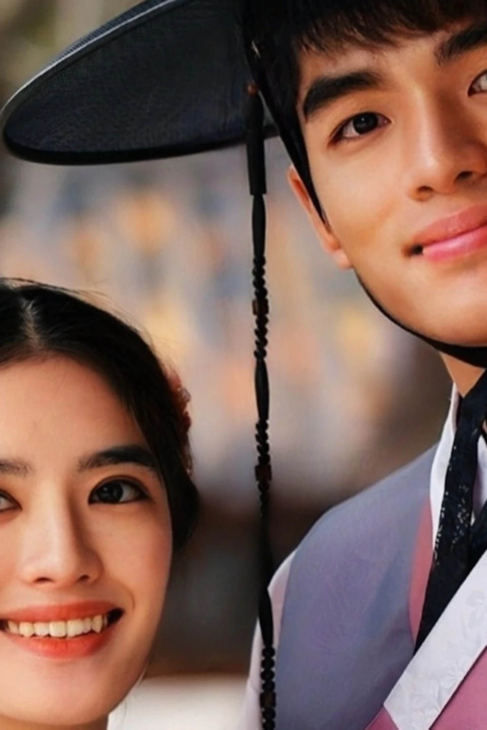 A couple in traditional Korean attire poses outdoors with a historic building and trees in the background.