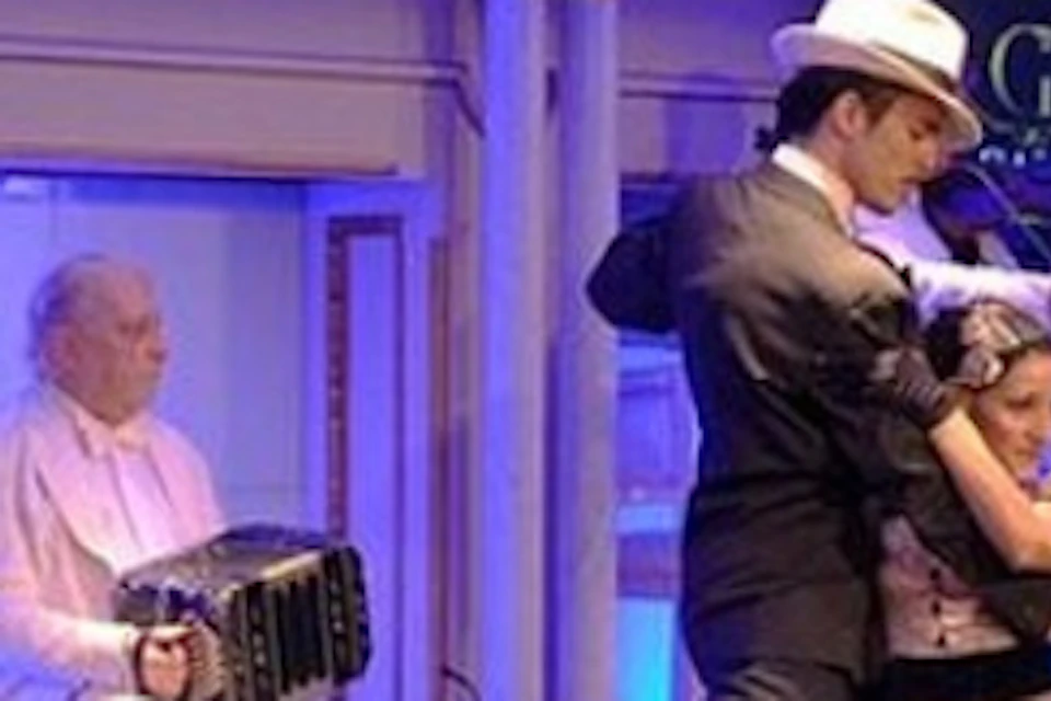 A couple performs a tango on stage with a musician playing the bandoneon in the background. Other musicians are partially visible.